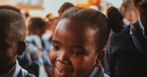 Uma menina jovem sorrindo para a câmera com outras crianças ao fundo.